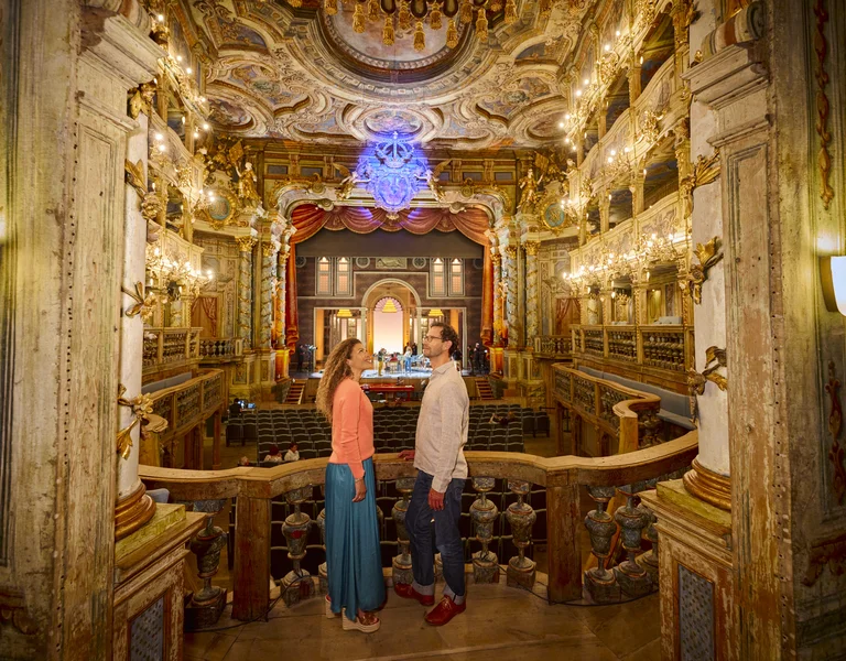 Two people stand facing each other in an ornate, golden theater balcony.