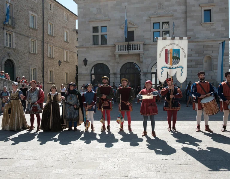 Group of people in medieval costumes standing in a town square with drums and a large banner.