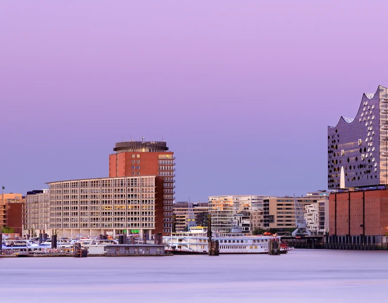 City skyline at sunset with modern buildings and a purple sky reflecting on the water.