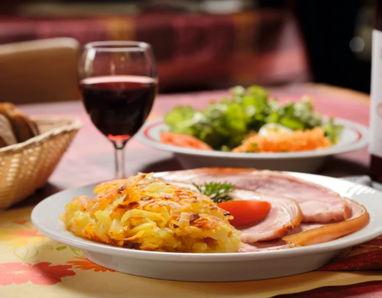 Ham, rösti, and a tomato slice on a plate with a glass of red wine and salad.