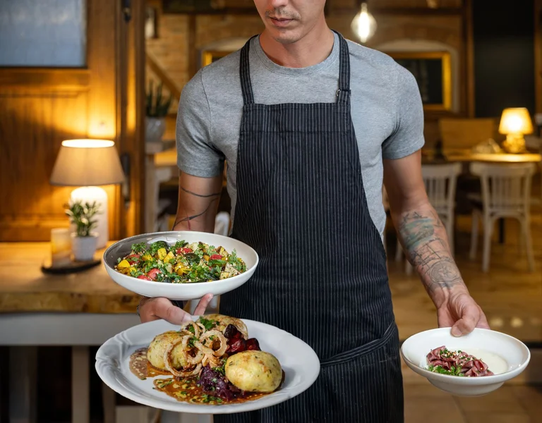 Waiter in an apron carrying three plates of food in a cozy restaurant.