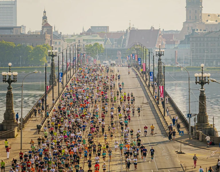 A large crowd of runners crosses a city bridge during a marathon on a sunny day.