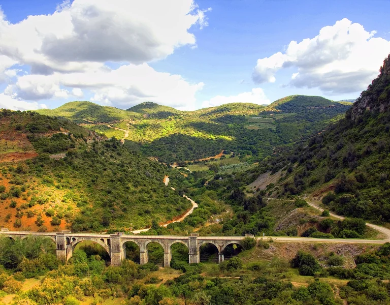 Stone bridge crosses a lush green valley with hills under a partly cloudy sky.