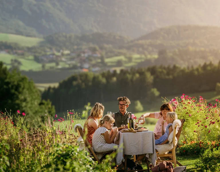 Four people enjoying a meal at a table in a scenic, sunlit mountain meadow.