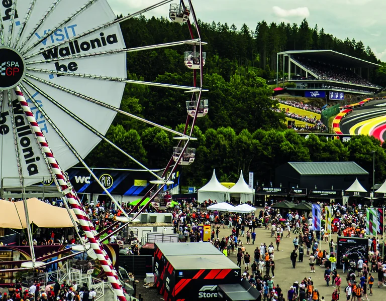 Large crowd at a racing event with a Ferris wheel and winding racetrack in the background.