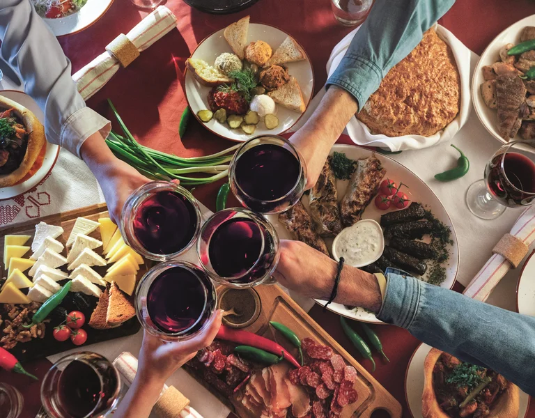 Four people toasting with red wine over a table filled with assorted food dishes.