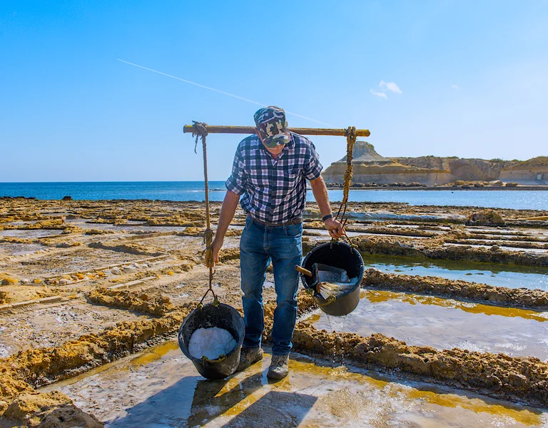 Man carrying buckets of salt near coastal salt pans under a clear blue sky.