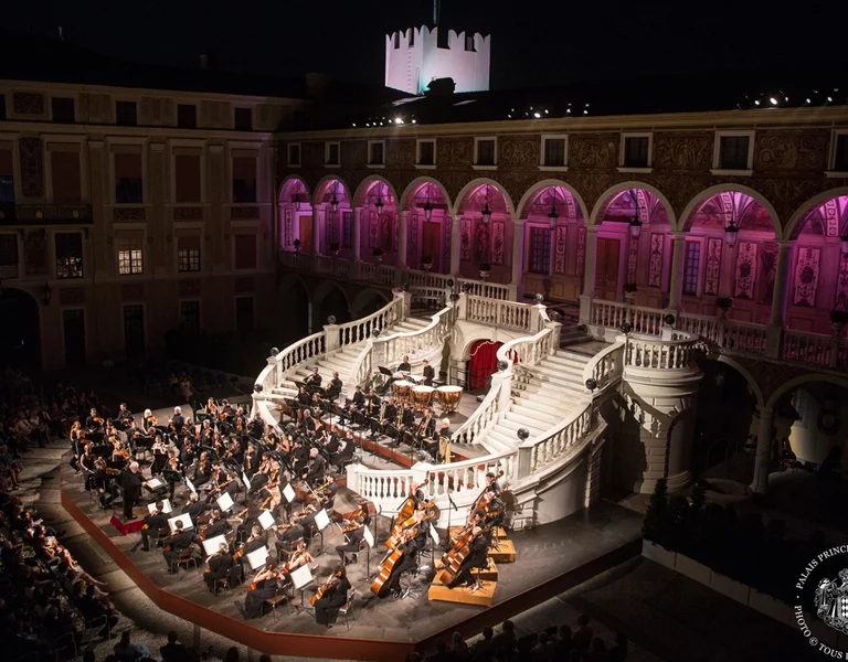 Outdoor orchestra performing on an ornate stage at night, surrounded by historic architecture.
