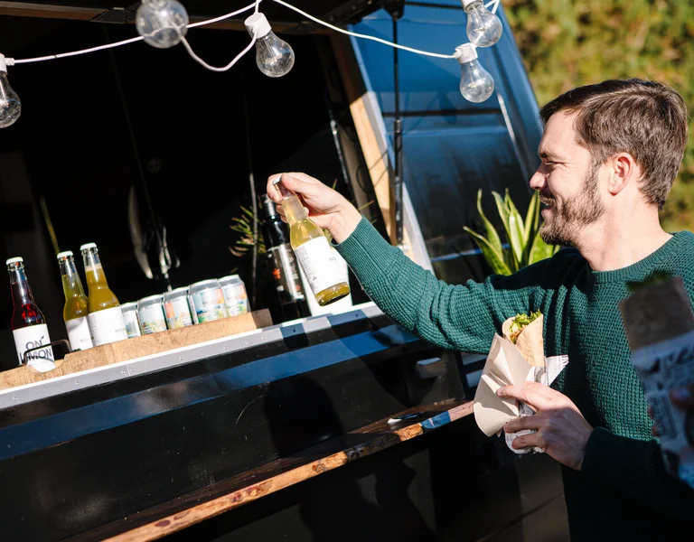 Man at a food truck selecting a drink, holding food, with string lights above.