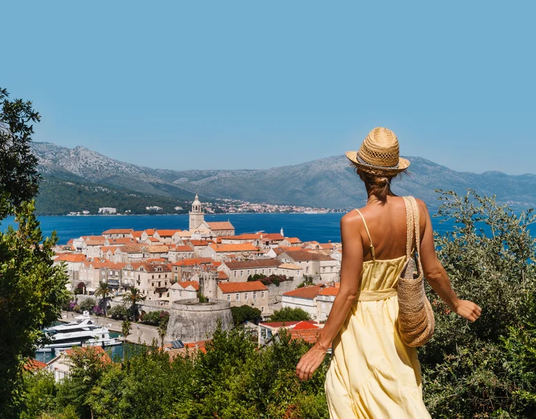 Woman in a yellow dress overlooks a coastal town with red rooftops and blue sea.
