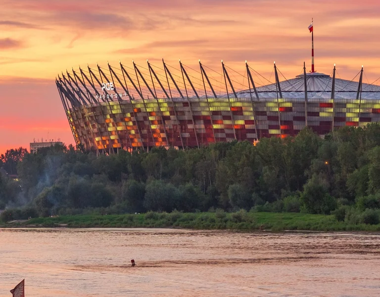 Stadium behind trees at sunset, river in the foreground, sky with orange and pink hues.