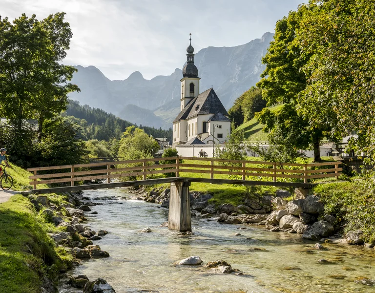 Wooden bridge over a clear stream, church with mountains in the background, lush green trees.