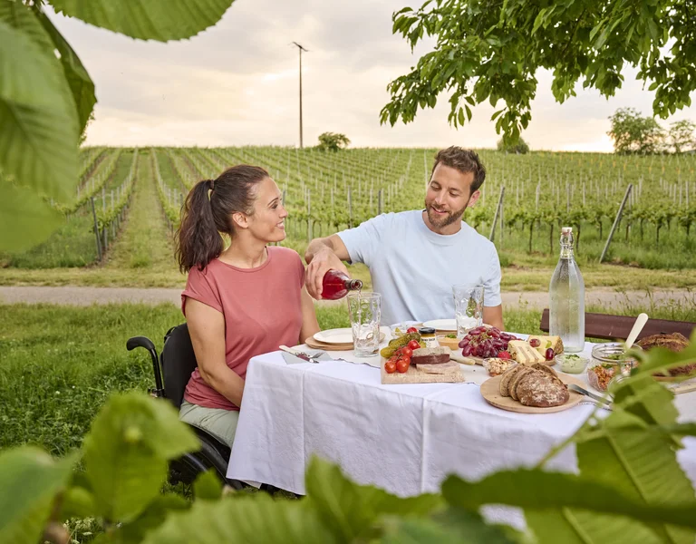 A couple shares a meal at an outdoor table in a vineyard, surrounded by greenery.