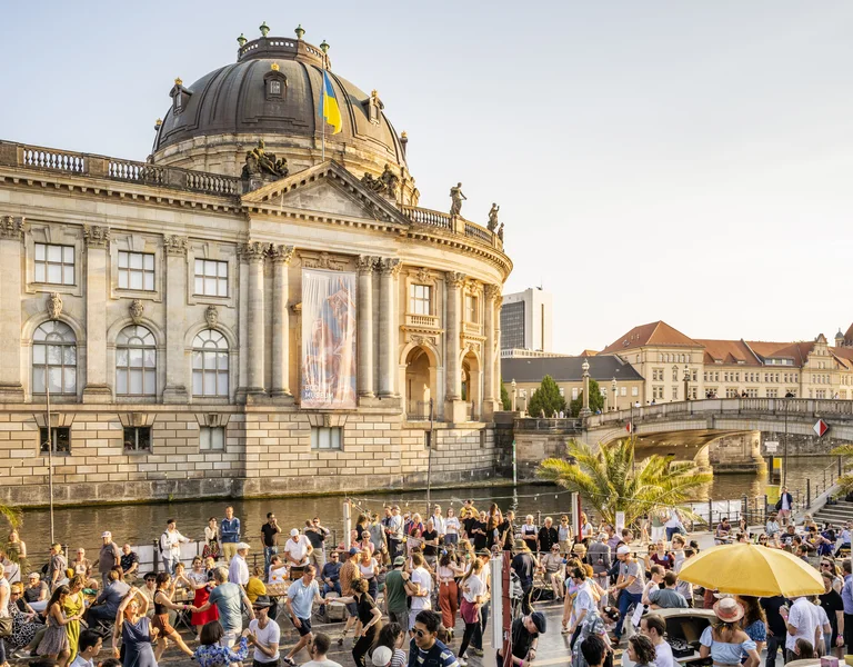 Large crowd gathers by a historic domed building near a river on a sunny day.