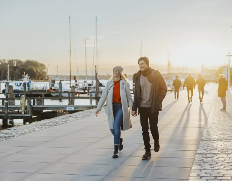 Couple walking along a marina at sunset, smiling, with boats in the background.
