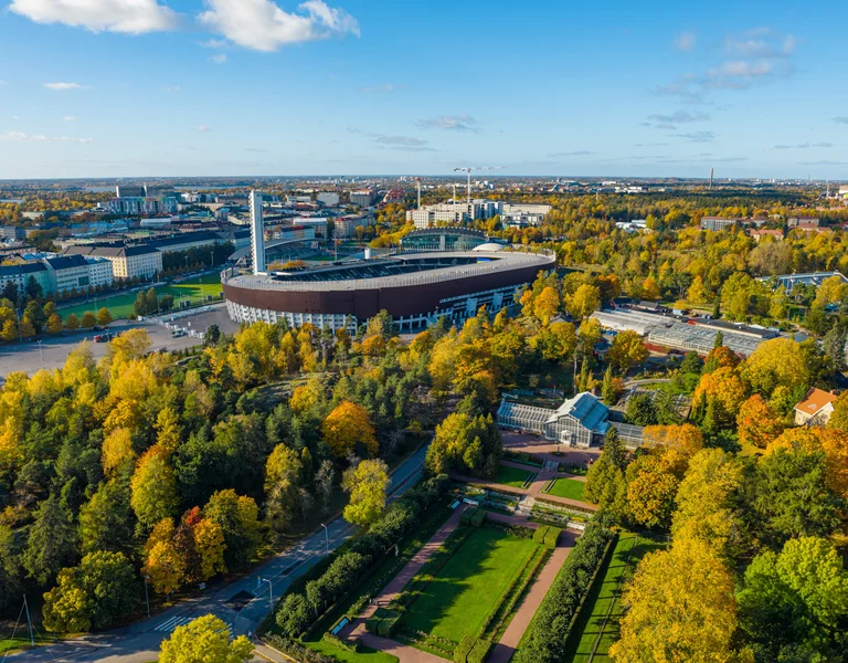 Aerial view of a stadium surrounded by autumn trees and city buildings under a blue sky.
