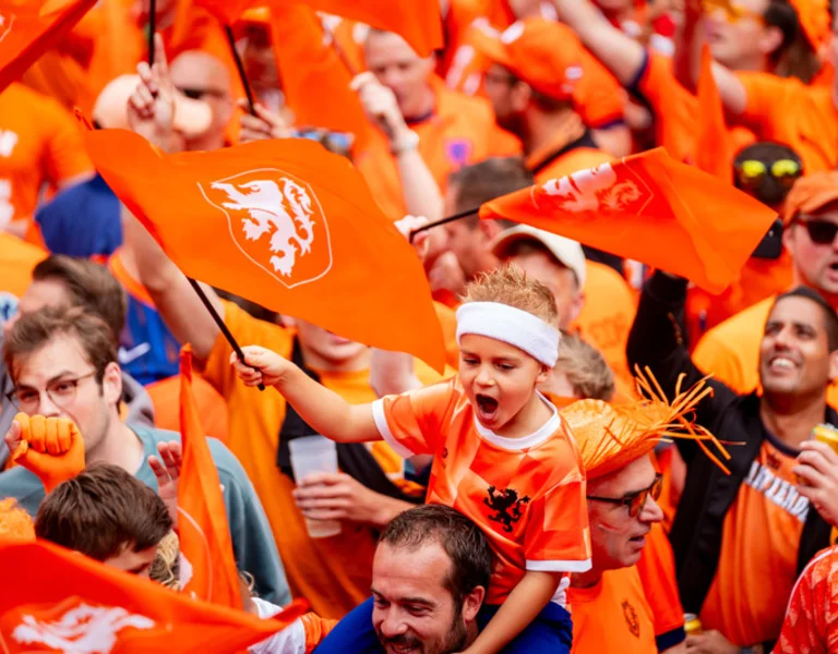 Crowd of Dutch soccer fans in orange, waving flags and cheering enthusiastically.