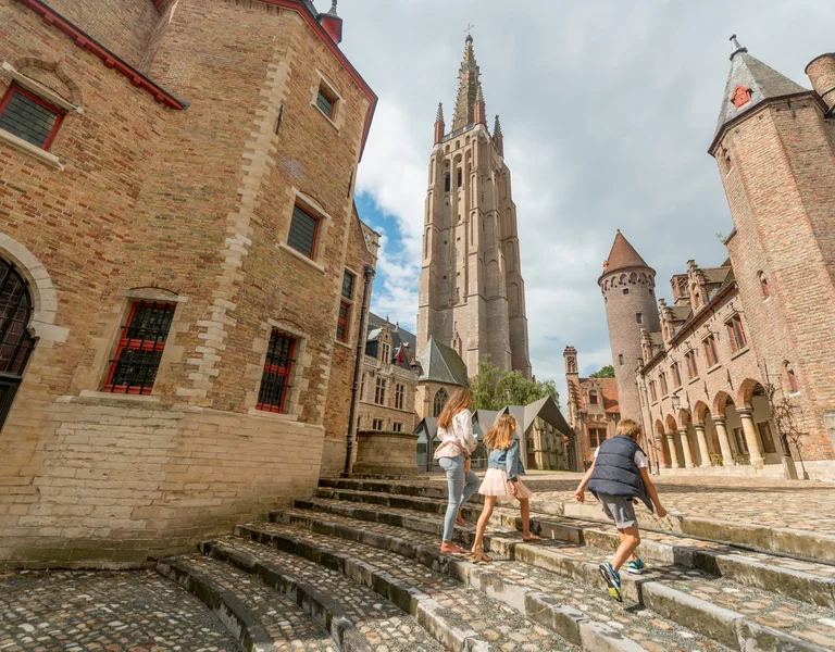 Three people walk up stone steps in a medieval European city with a tall church tower.