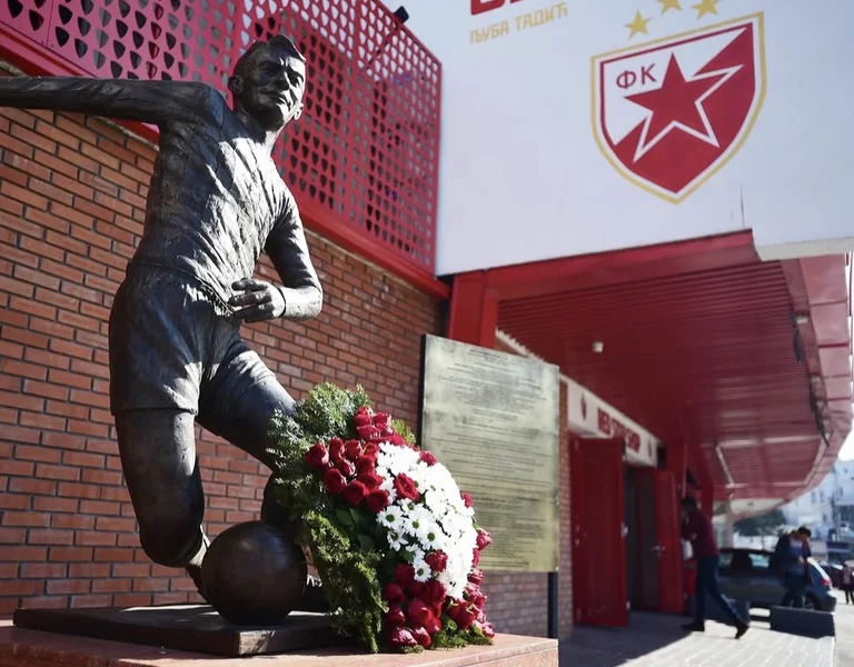 Bronze soccer player statue with floral wreath, stadium and red star logo in background.