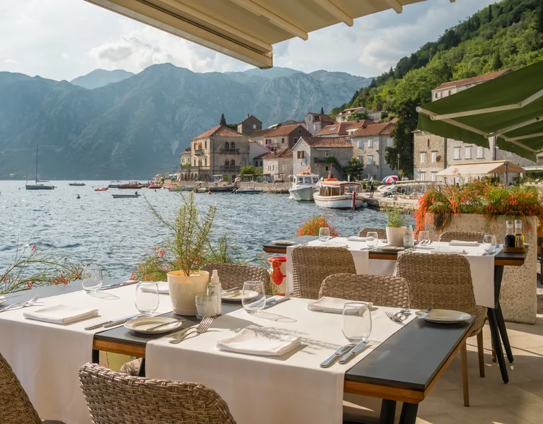 Outdoor restaurant by a calm lake, mountain backdrop, and village buildings in sunlight.