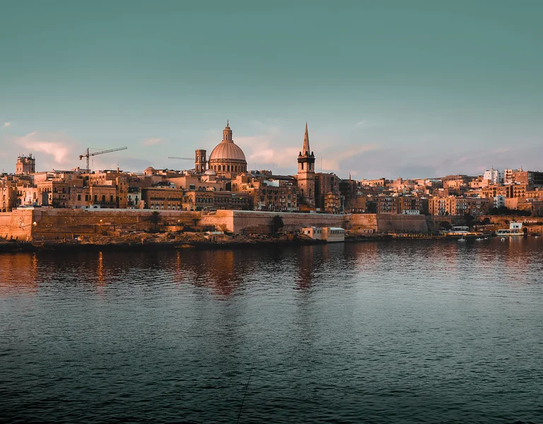 Old city skyline with domed church and spire reflected on calm waterfront at sunset.