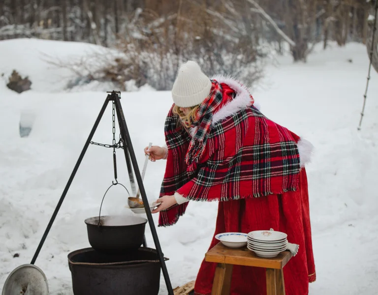 Person in red plaid cloak cooking over an outdoor cauldron in snowy winter scenery.