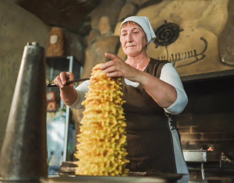 Woman in traditional clothing preparing a tall twisted pastry in a rustic kitchen.
