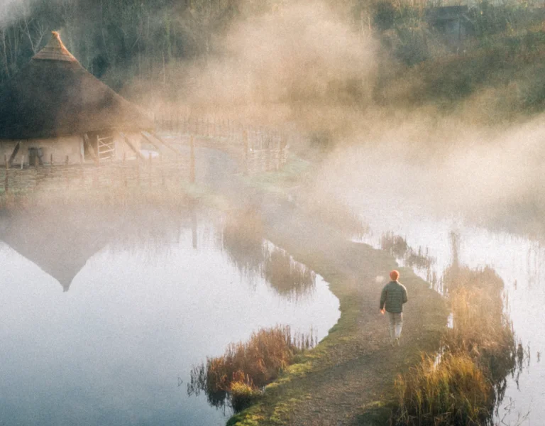 Foggy lakeside village with thatched cottages and autumn trees reflected in calm water.
