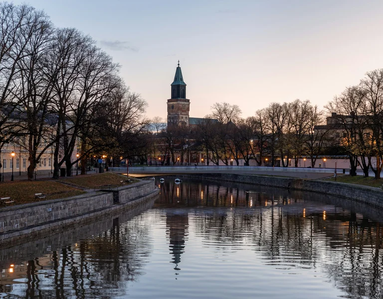 Calm river with stone banks, trees, and a tall church tower at dusk.