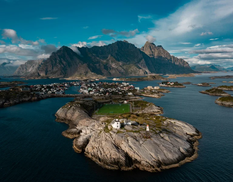 Rocky island with a lighthouse, surrounded by blue sea and distant mountains under a cloudy sky.