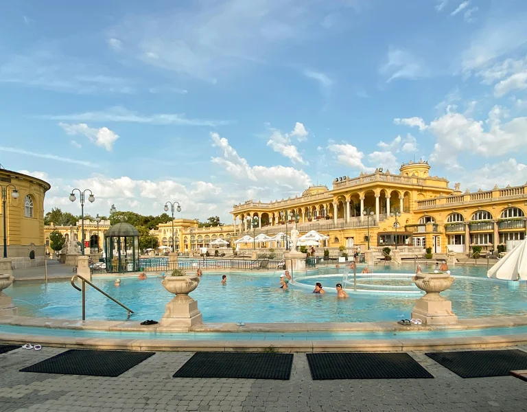 Outdoor thermal pool with people swimming, surrounded by ornate yellow historic buildings.