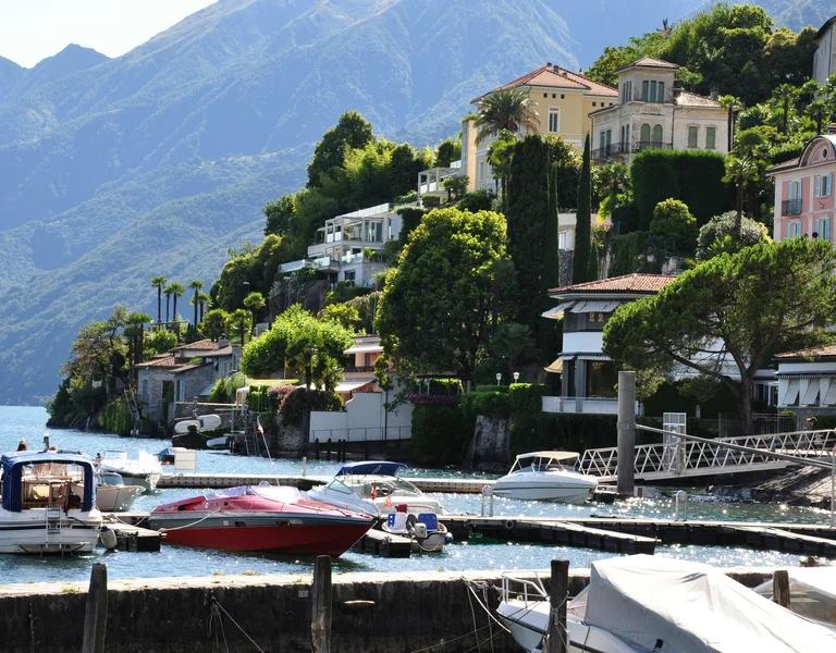Colorful lakeside houses with boats docked, mountains and greenery in the background.