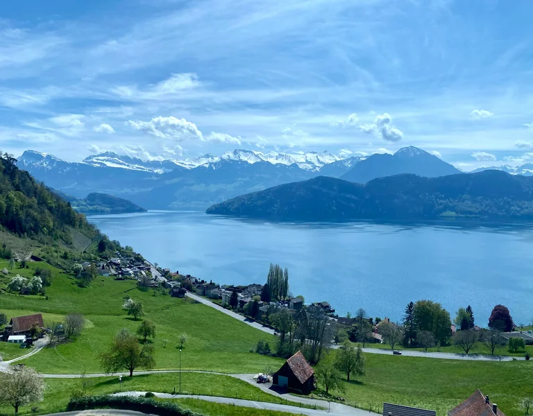 Green hills and scattered houses overlook a lake with mountains in the background under a blue sky.
