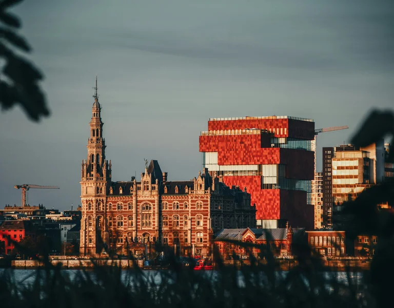 Historic building and modern red block structure by the water at sunset.