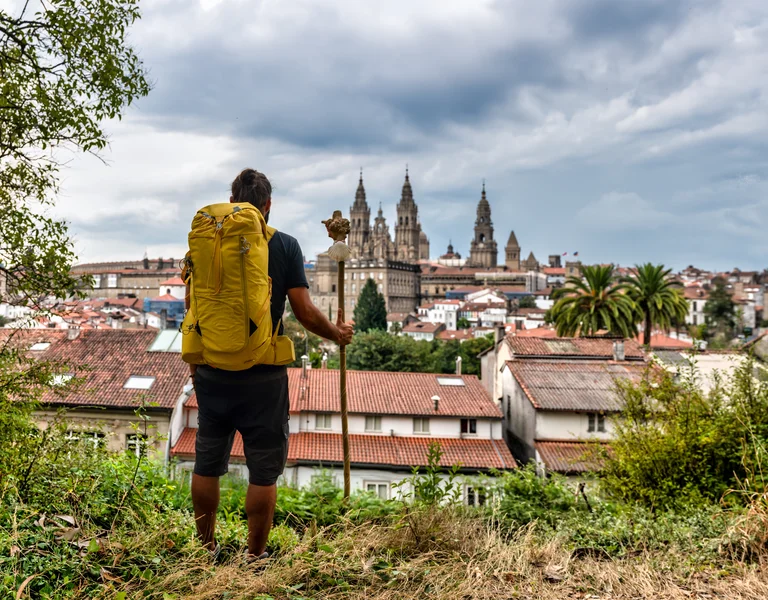 Hiker with yellow backpack overlooking a city with church spires under cloudy sky.