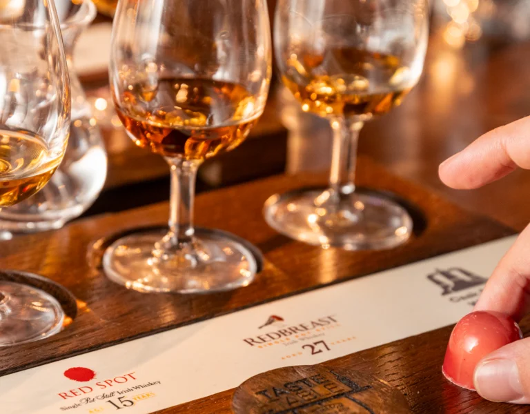 Hand reaching towards tasting flight of whiskey glasses on a wooden tray.