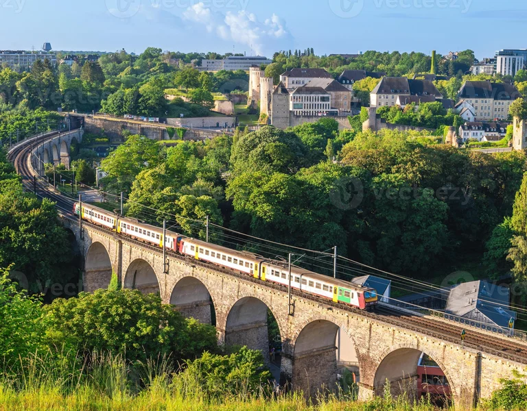 Passenger train crossing a stone viaduct surrounded by lush greenery and historic buildings.