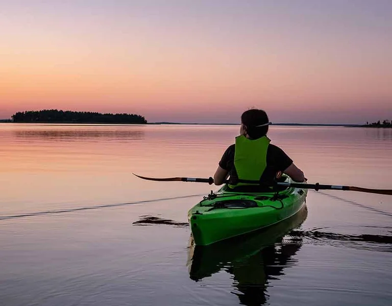 Person kayaking on calm water at sunset with islands in the distance.