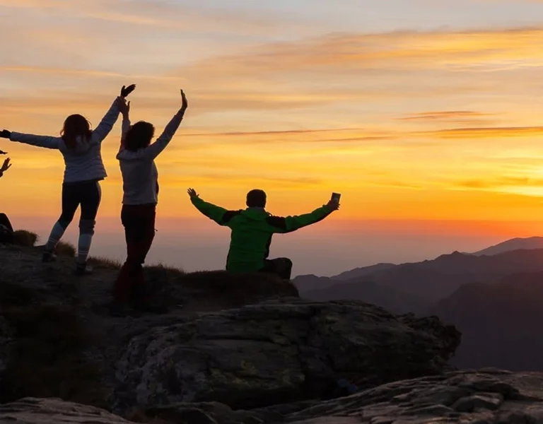 Group of people on a mountain peak celebrating at sunset with arms raised.