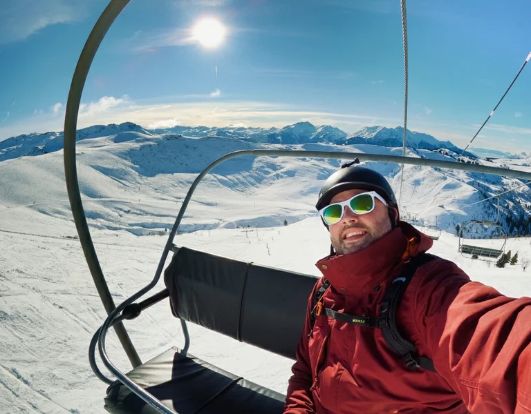 Man in sunglasses and red jacket on a ski lift with snowy mountains in the background.