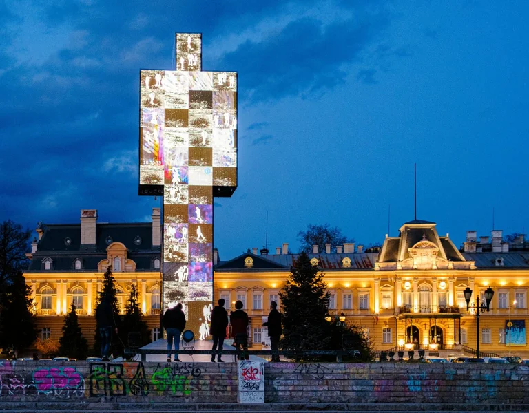 Illuminated cross monument with photo projections, people standing in front, buildings lit at dusk.