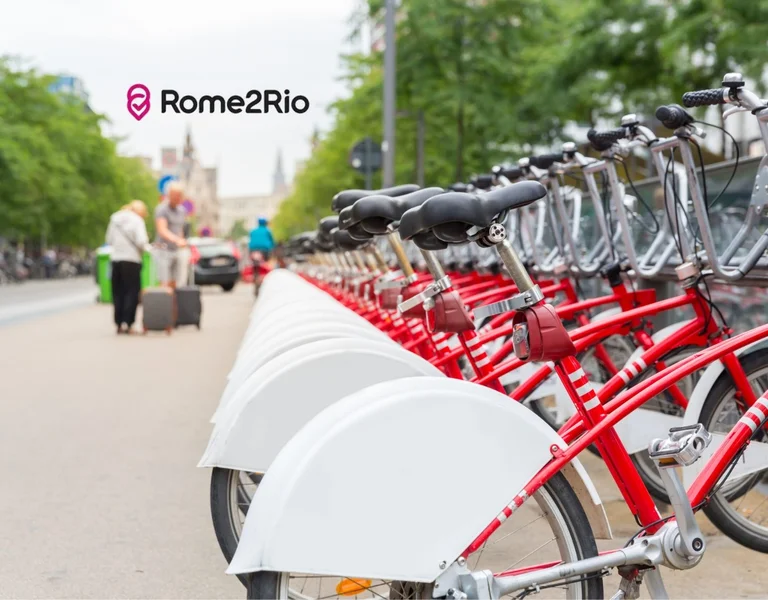Row of red rental bikes on a city street with people and trees in the background.