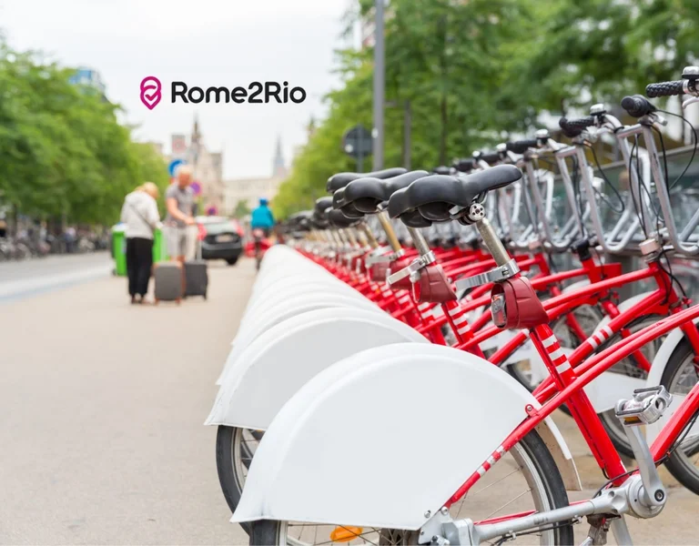Linha de bicicletas vermelhas para aluguel em uma rua da cidade com pessoas e árvores ao fundo.