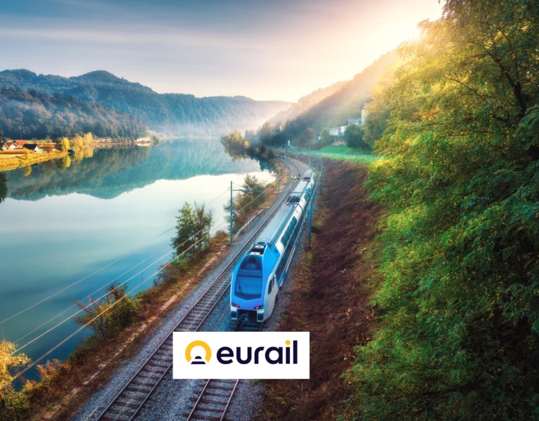 A blue train travels by a lake and forested hills at sunrise, with the Eurail logo displayed.