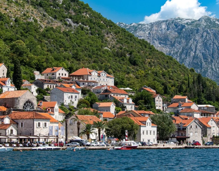 Coastal village with red-roofed buildings, a church tower, and mountains in the background.