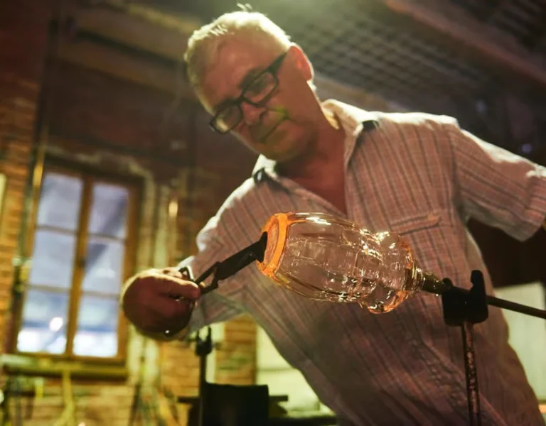 Man shaping molten glass with tools in a workshop.