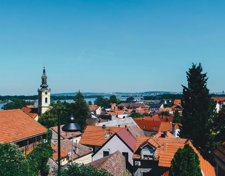 Town with red rooftops, church steeple, trees, and river under a clear blue sky.