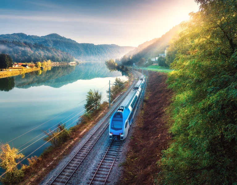 Modern train traveling beside a calm river, surrounded by trees and hills at sunrise.