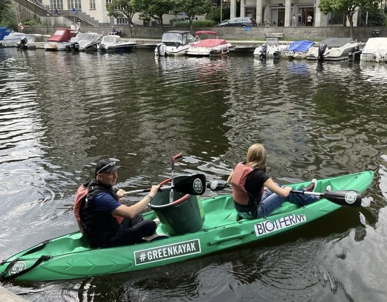 Two people in a green kayak collecting trash from a canal near docked boats.