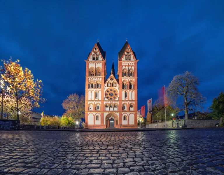 Illuminated cathedral with twin towers at dusk, viewed from a cobblestone square.